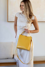 Load image into Gallery viewer, Lady wearing a long white dress holds a yellow vegan leather shoulder bag.
