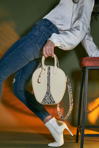 Cream and snake print vegan leather oval round shoulder handbag with silver hardware is held against the bent leg of a women wearing blue denim jeans and a white shirt.